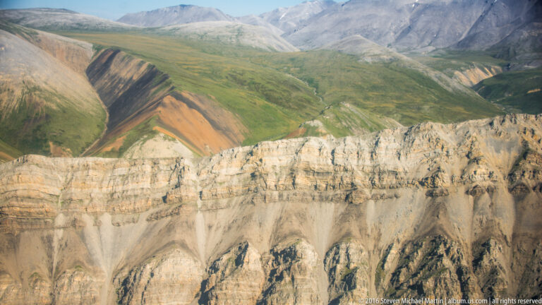 A pale layered escarpment with vertical gullies below rust-colored mountain slopes.