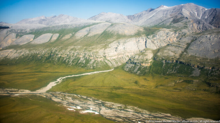 A braided river with pale gravel islands curves across a broad green valley.