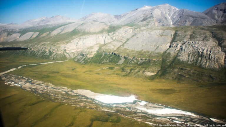 A braided river crosses a green valley below a pale, banded mountain slope.