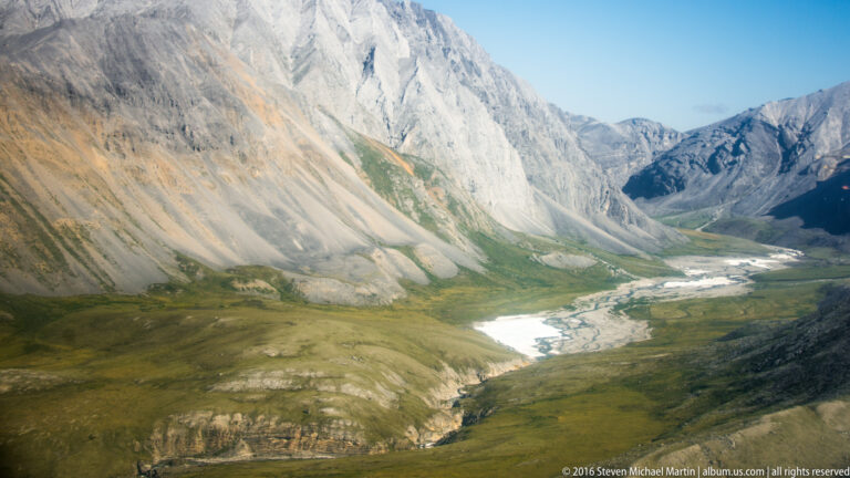 A broad mountain valley with a braided stream and snow patches on steep slopes.