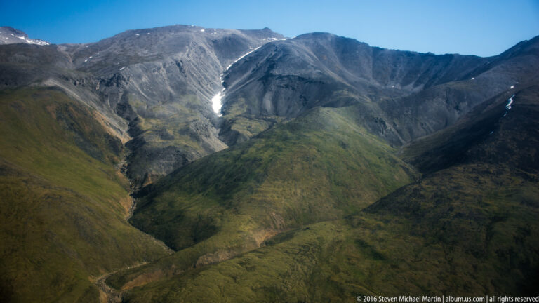 A green ravine runs between rounded ridges toward a small snow patch on a distant slope.
