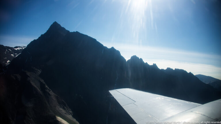 An airplane wing points toward a dark mountain ridge silhouetted against bright sunlit sky.