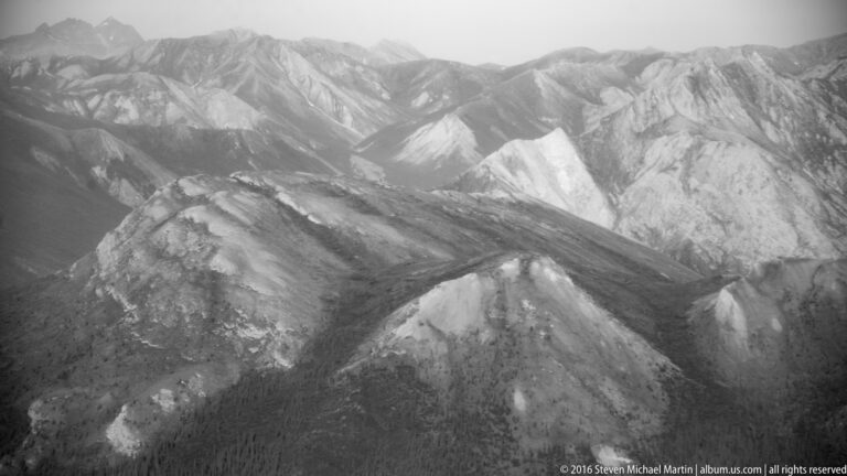 Pale rounded ridges descend into dark valleys, with sharper peaks and haze beyond in a monochrome aerial view.