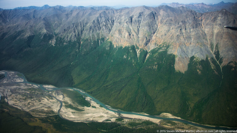 A braided river bends below a steep mountain wall with sunlit rock near its summit.