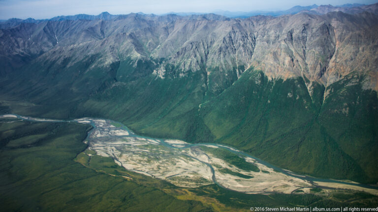 A broad braided river fan spreads across a valley below steep green mountains.