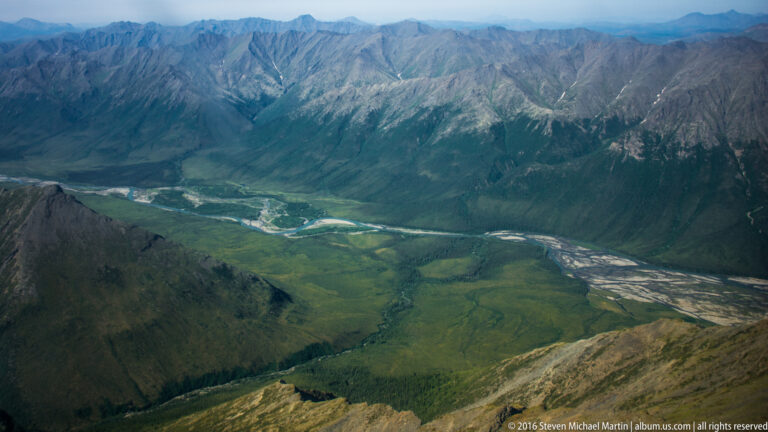 Pale braided river channels divide around gravel bars below a green mountain valley.