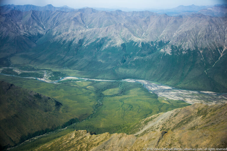 A braided river crosses a green valley floor between steep mountain slopes.