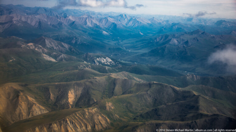 Aerial panorama of layered Arctic mountain ridges beneath a cloudy horizon, with cloud crossing the right side.