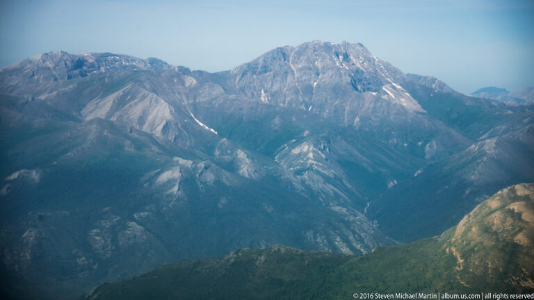 A pale summit rises above blue, hazy mountain ranges and shadowed valleys, with a green-brown slope in the foreground.