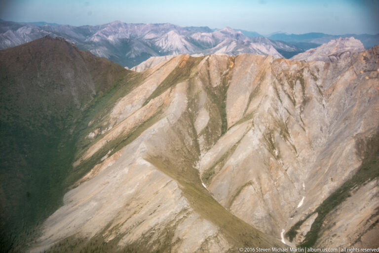 A sunlit pale ridge angles across the frame above a dark green valley and layered mountains.