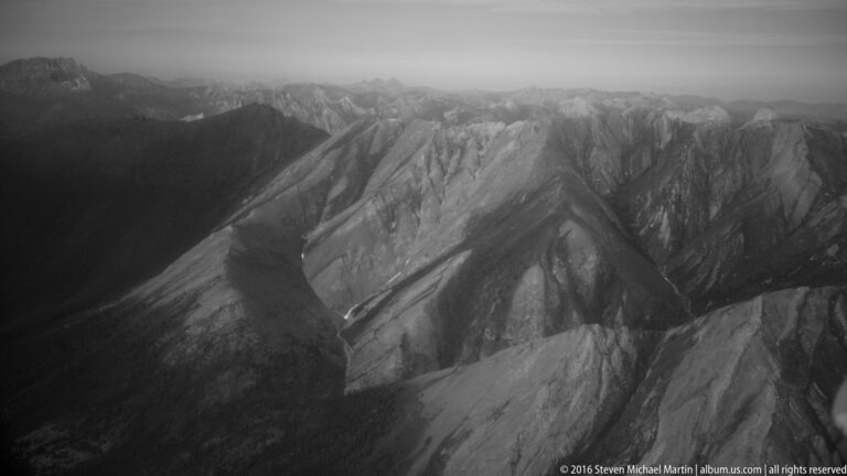 Bright mountain ridges alternate with dark troughs in a black-and-white aerial view fading into haze.
