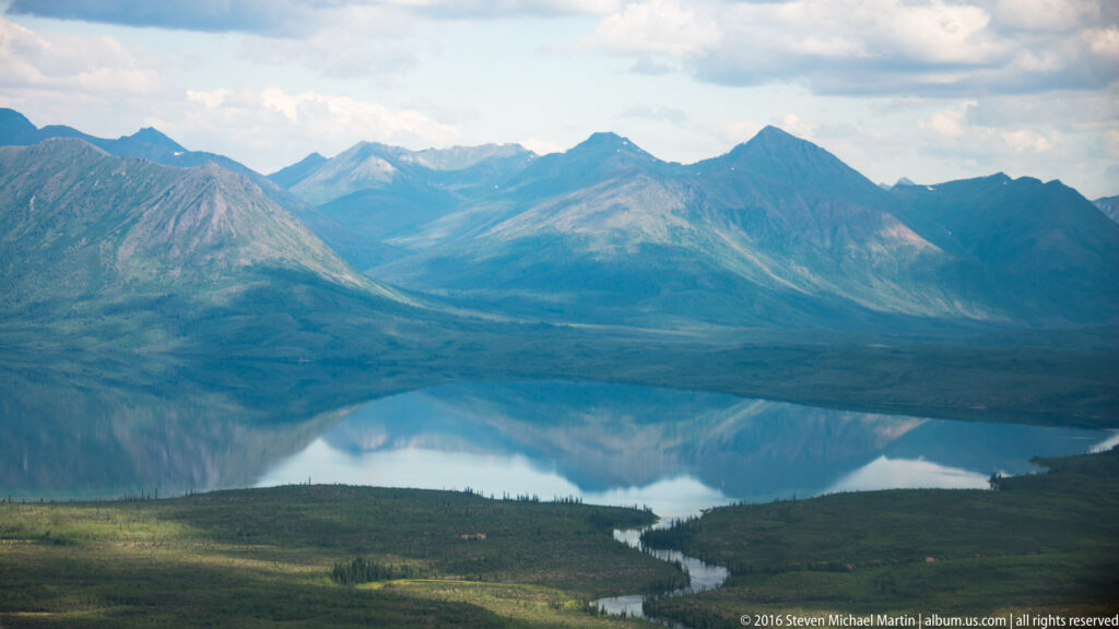 Aerial view of a winding river entering a reflective lake below steep, treeless Alaska Range peaks.