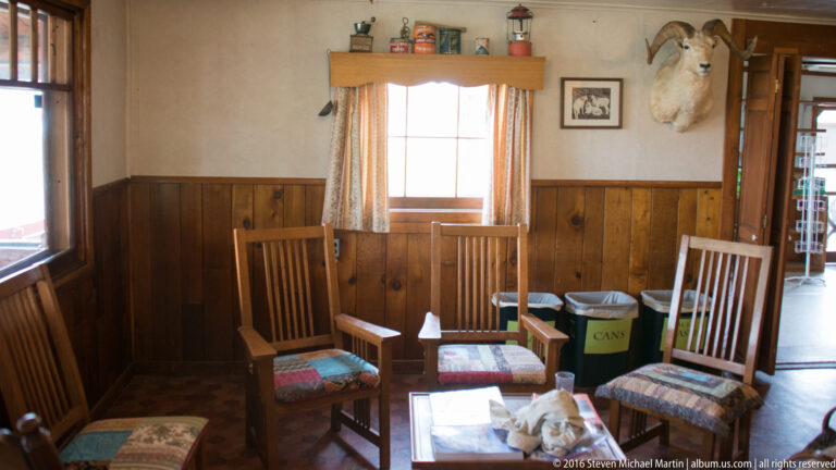 Wooden chairs and small tables sit beneath a mounted moose head in a lodge room with dark wainscoting.