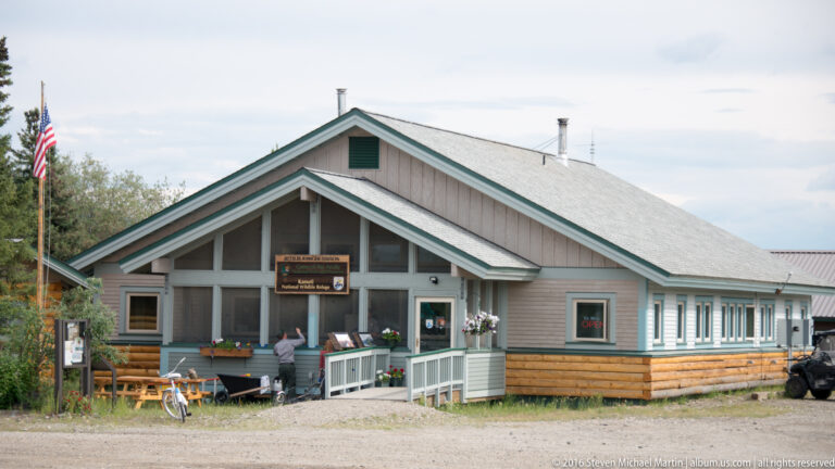 A low gabled building has a covered porch, long windowed wing, log-clad base, and an American flag.