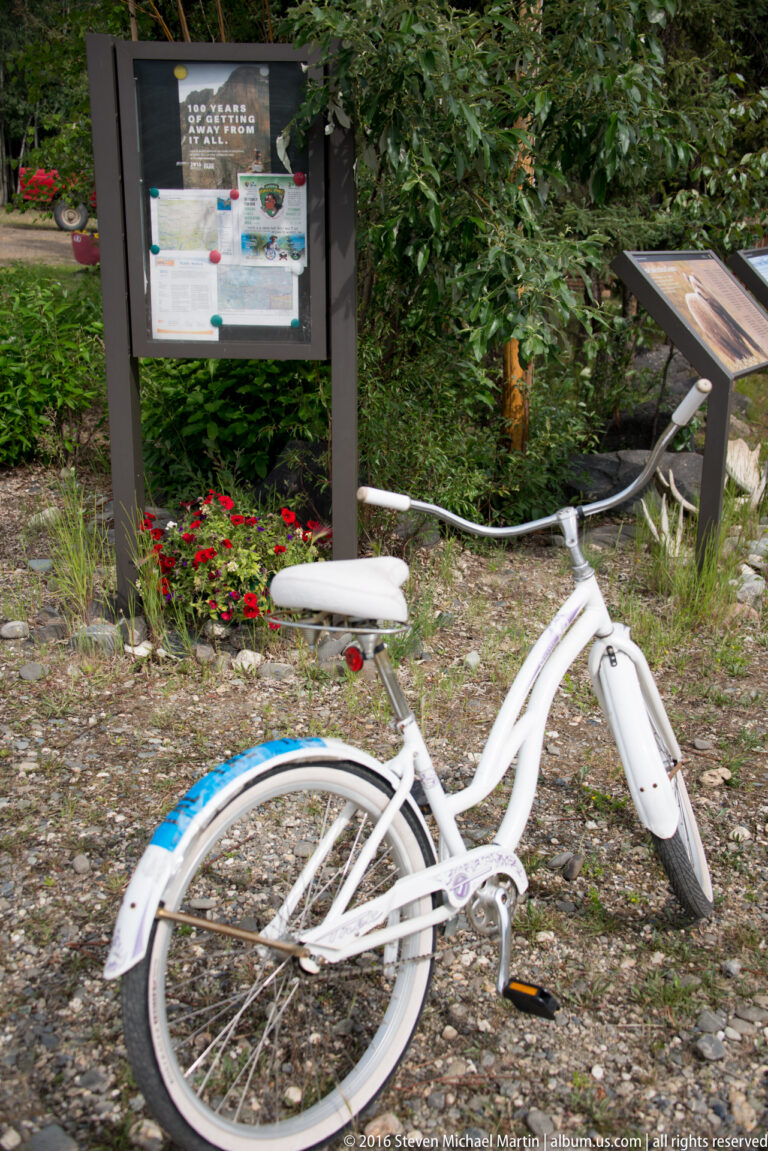 A white bicycle stands beside an outdoor information board and angled interpretive panel among shrubs and flowers.