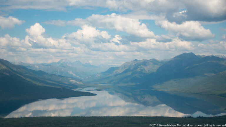 Clouds and mountain ridges reflect in a long lake seen from above, with smaller lakes continuing into the distance.