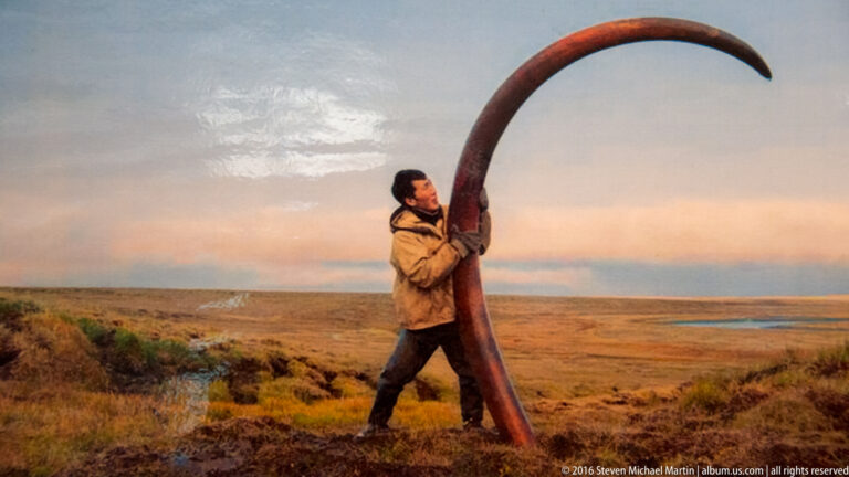 A man carries a tall, curved tusk-like object across open tundra beneath a pale evening sky.