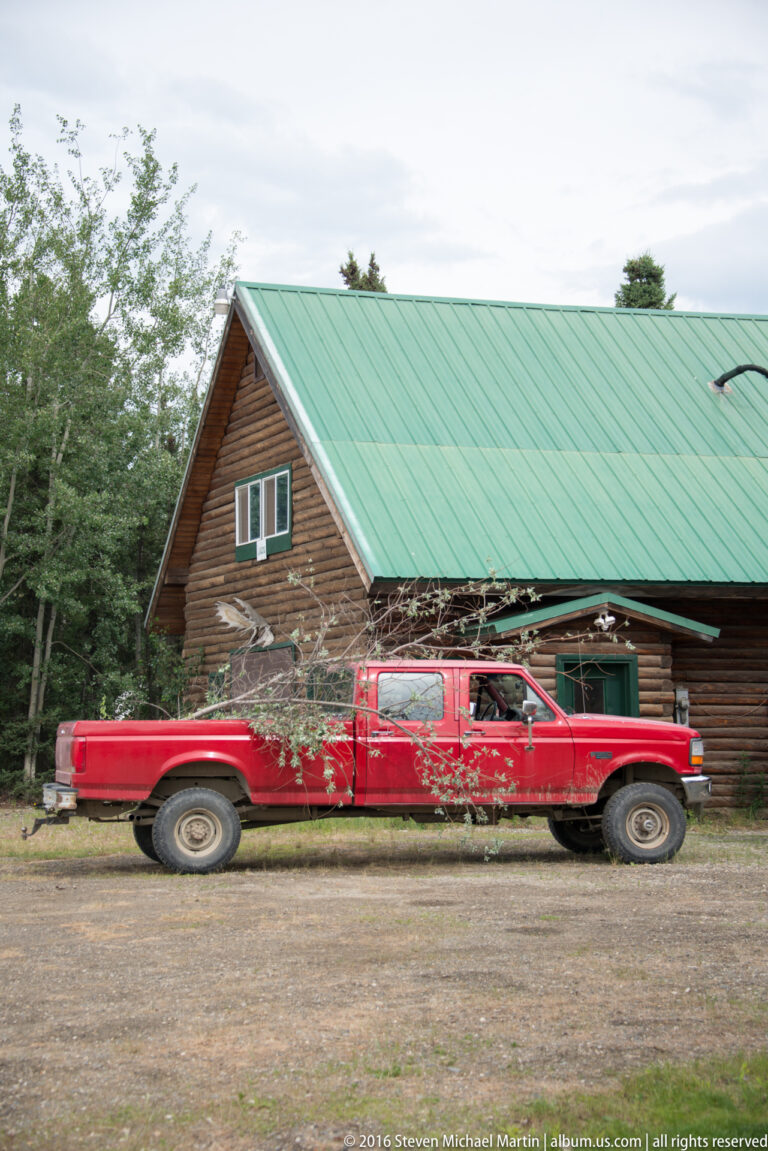 A red pickup with leafy branches across it is parked beside a log cabin with a green metal roof.