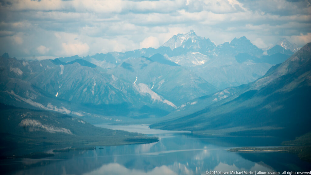 A blue lake reflects rugged, partly snow-streaked peaks within a steep Alaska mountain valley.