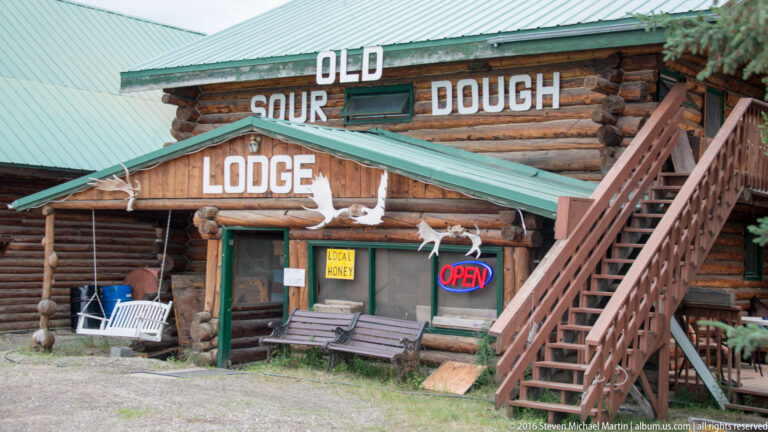 The Old Sour Dough Lodge has large white lettering, an exterior staircase, mounted antlers, and an open sign.