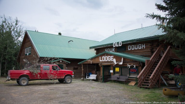 Log lodge with a green roof and red pickup parked outside on a gravel forecourt.