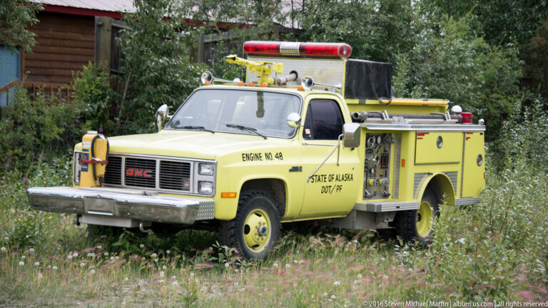 Yellow GMC fire engine with roof lights and side compartments parked in tall grass and wildflowers.