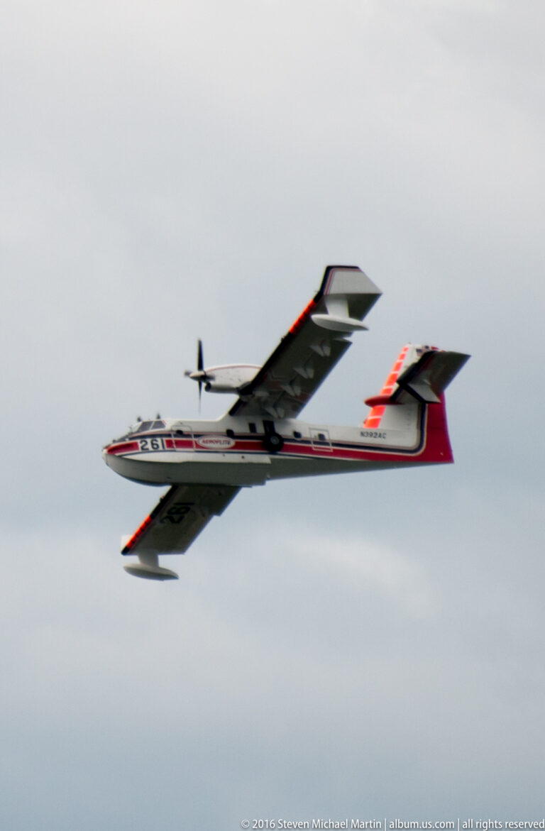 Small red, white, and dark propeller airplane flying across a pale cloudy sky.