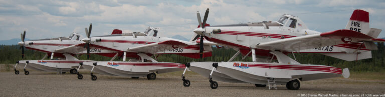A row of red-and-white high-wing utility airplanes parked on a gravel airfield.