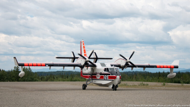 Front-facing red-and-white twin-engine high-wing airplane on a gravel strip beneath low clouds.
