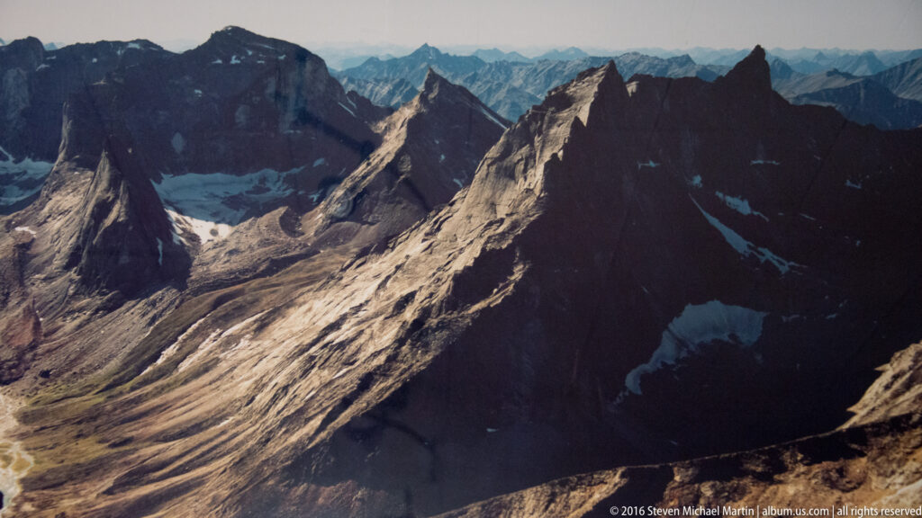 Aerial view across sharp mountain ridges, shadowed valleys, snow patches, and small lakes.