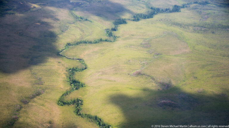 A narrow stream meanders through open green tundra, with shrubs tracing parts of its banks.