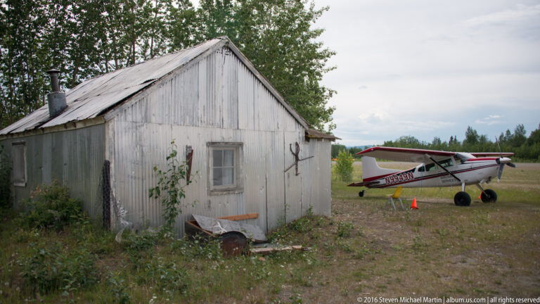 Small red-and-white airplane parked beside a weathered gray shed at a rural airfield.
