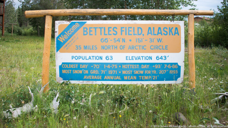 Blue-and-yellow Bettles Field sign in grass, listing coordinates, elevation, population, and climate records.