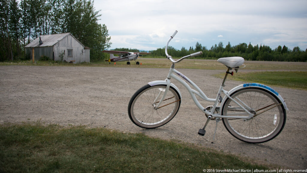 White step-through bicycle on gravel, with an airplane and shed in the background.
