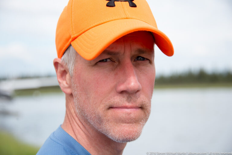 A man in a bright orange cap and blue shirt looks toward the camera beside a blurred waterway.