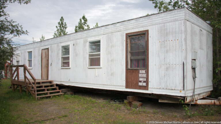 White rectangular bunkhouse on short supports, with a small porch and a BUNK HOUSE sign on the door.