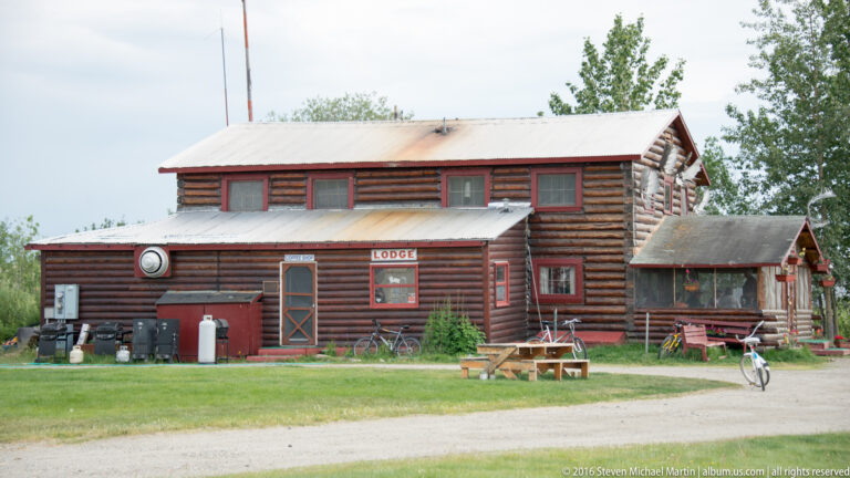 Side view of a two-story log lodge with red-trimmed windows, utility equipment, and a bicycle.
