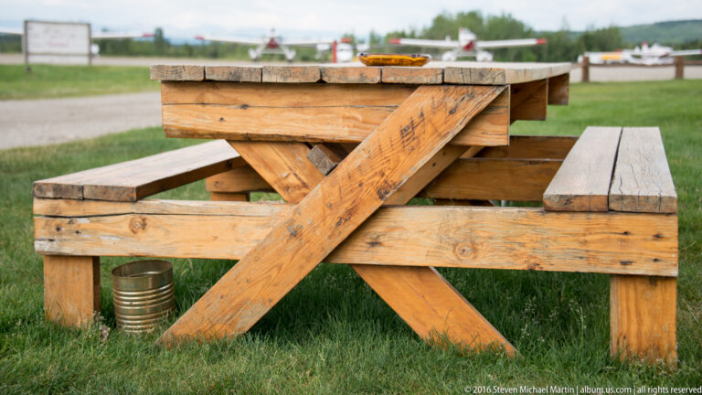 Weathered wooden picnic table at an airfield, with parked airplanes blurred in the distance.