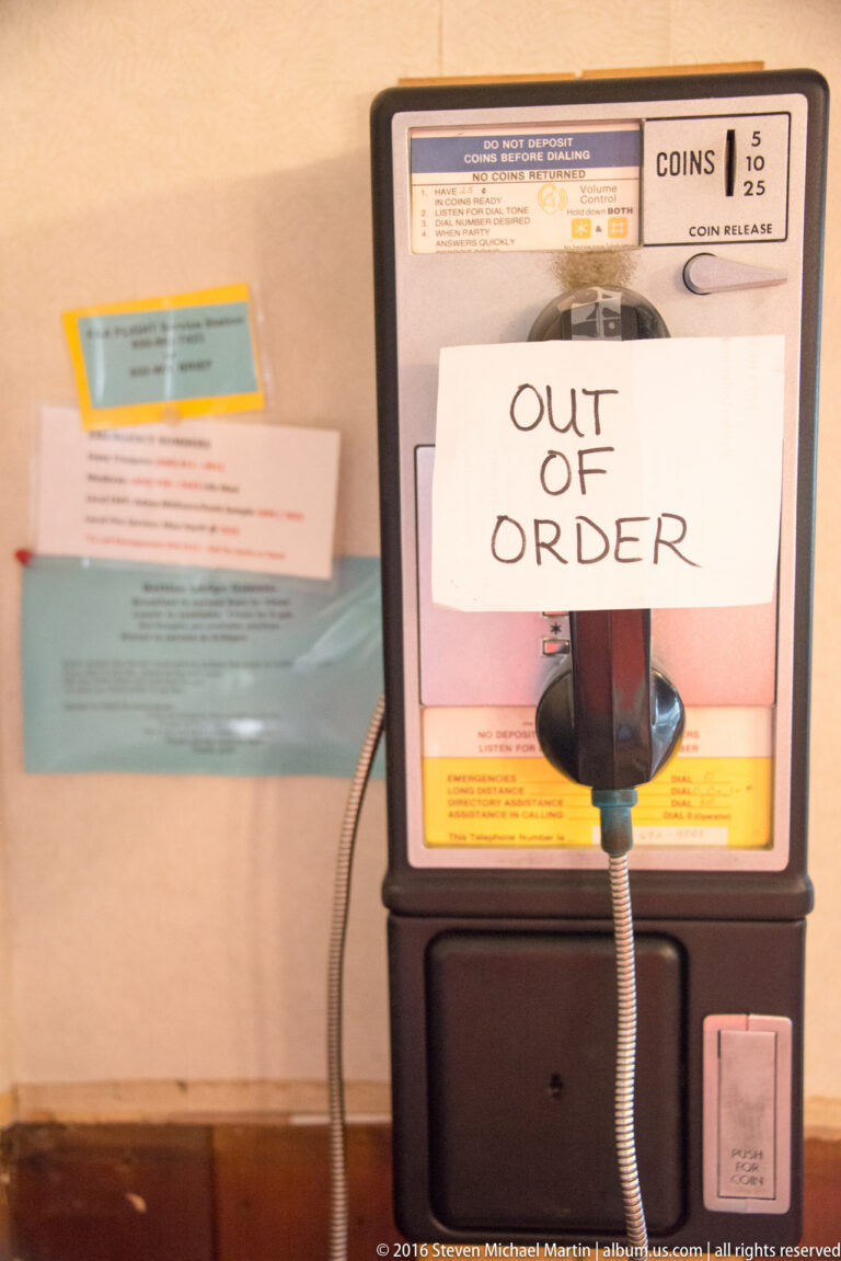 A handwritten out-of-order notice covers a public telephone, with coin slots, instructions, handset, and cord visible.
