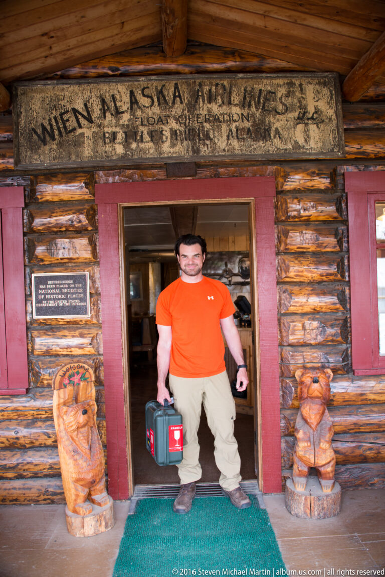 A man holding a case stands in a log building doorway between two carved wooden figures beneath a weathered aviation sign.