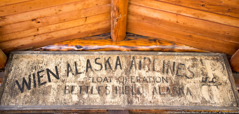 A faded wooden aviation sign under a timber roof names Wien Alaska Airlines and Bettles Field, Alaska.