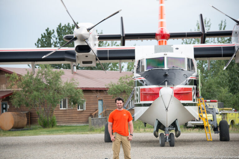 A man stands beside the nose of a parked three-propeller aircraft, with a ladder and timber buildings behind it.