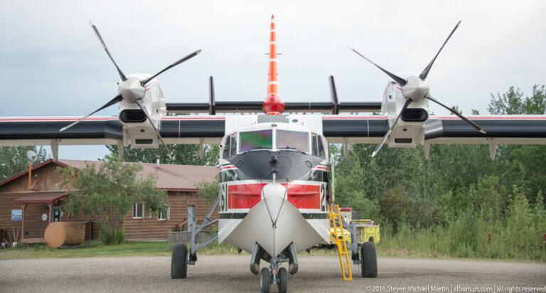 A three-propeller aircraft faces the camera at a small airfield, with a timber building and equipment behind it.