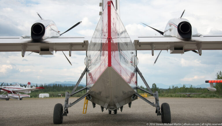 A low rear view shows an aircraft’s underside, landing gear, broad wings, and two propeller assemblies on an airfield.