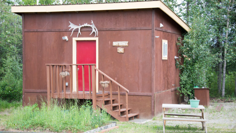 A small brown cabin with a red door and short porch stands among trees, with a white animal silhouette above the entrance.