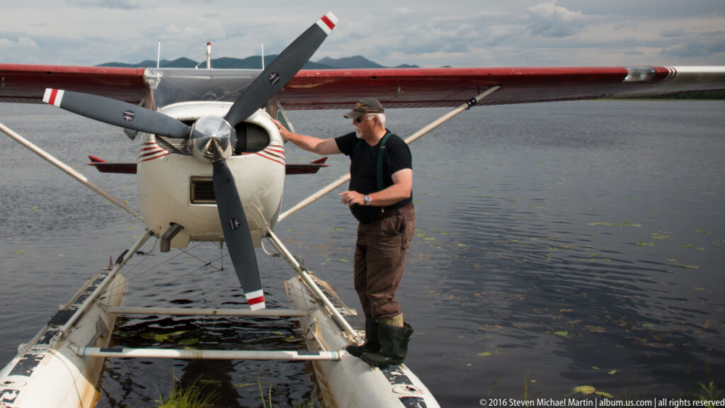 A man in waders stands on a floatplane pontoon beside the aircraft’s propeller on a calm river.