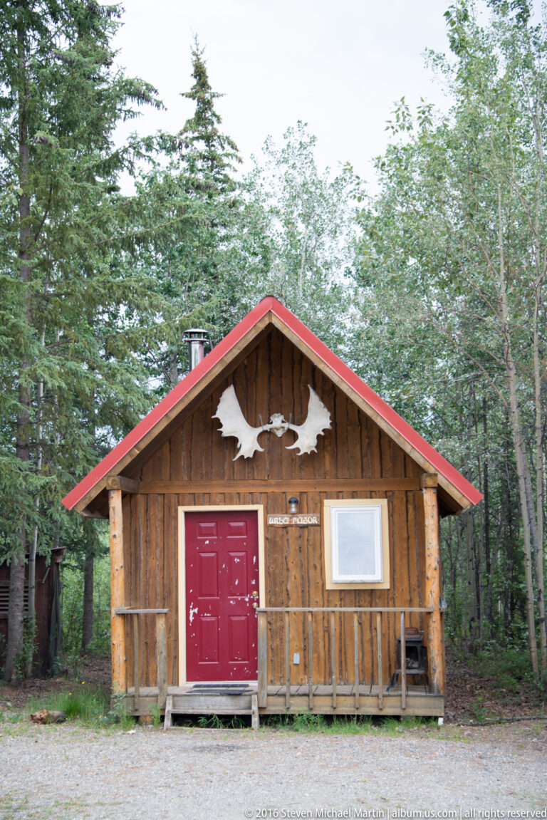 A log cabin with red double doors, front windows, and a pale antler-shaped ornament stands among trees.
