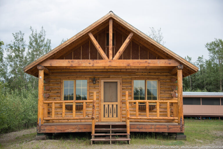 A log cabin has a deep covered porch, exposed timber gable trusses, a central door, and paired front windows.