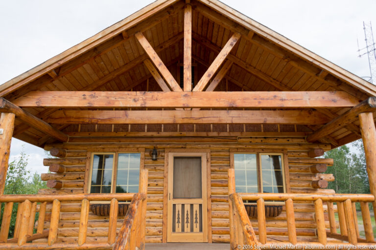 Exposed timber trusses and a broad crossbeam frame the underside of a log cabin’s porch gable.