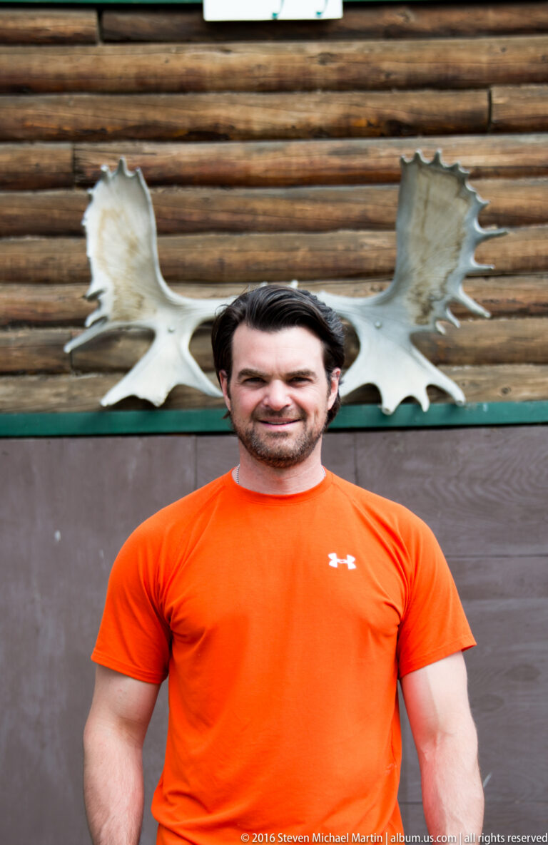 A man in an orange shirt poses in front of a large mounted moose-antler plaque on a log wall.